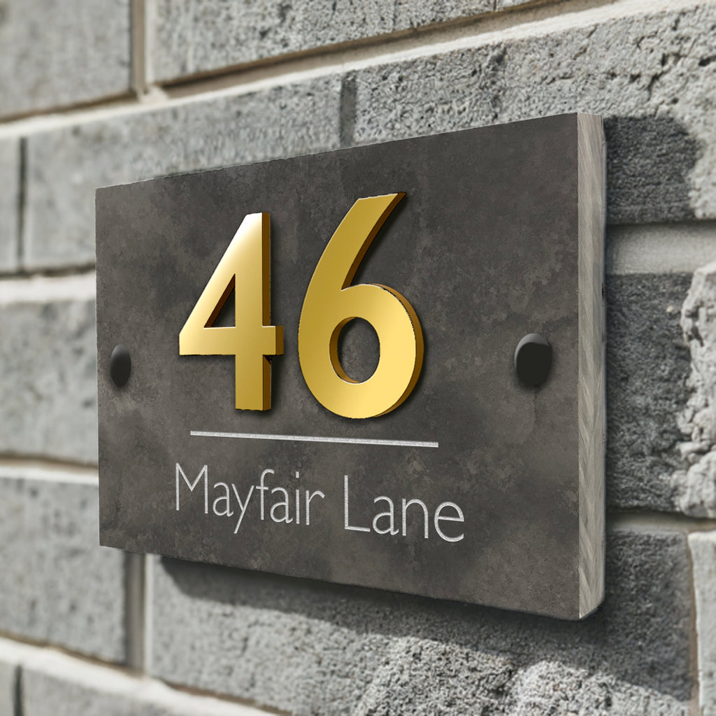 A house sign on a wall affixed with screws and finished with screw caps. The sign is constructed from 1cm thick honed slate and the number is gold acrylic material, giving a raised effect to the number. The street name "mayfair lane" is etched into the surace of the slate.