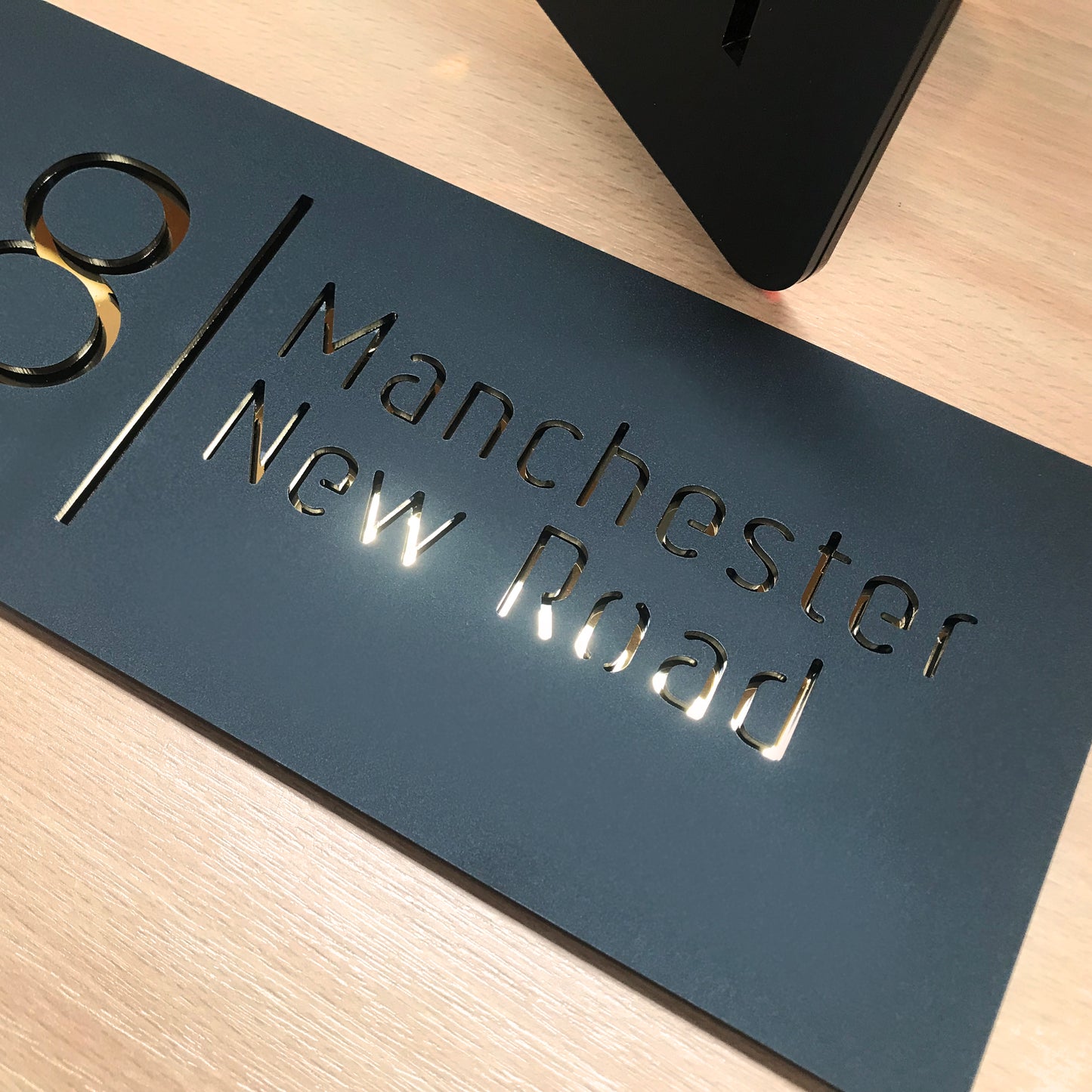 A contemporary stellano house sign. It is constructed from matt black acrylic, and the numbers and letters are laser cut from the material. A gold reflective mirror is bonded to the reverse allowing the viewer to see the numbers a reflective finish. The sign is rectangular and information on the sign consists of both house number and street name.