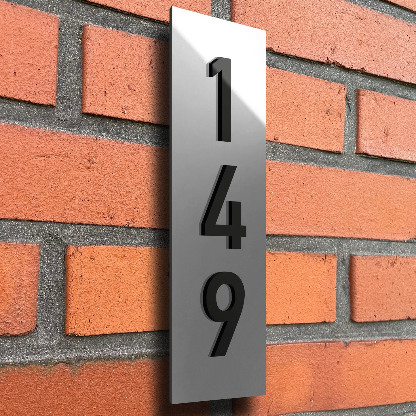 A house number sign made from metallic silver acrylic, the numbers are produced in glossy black material and are raised 3mm from the sigh surface to give a 3d effect. The sign is orientated vertically and the number '149' is also arranged vertically. The sign is affixed to an exterior house wall using a method designed to make the sign appear as if it is floating.