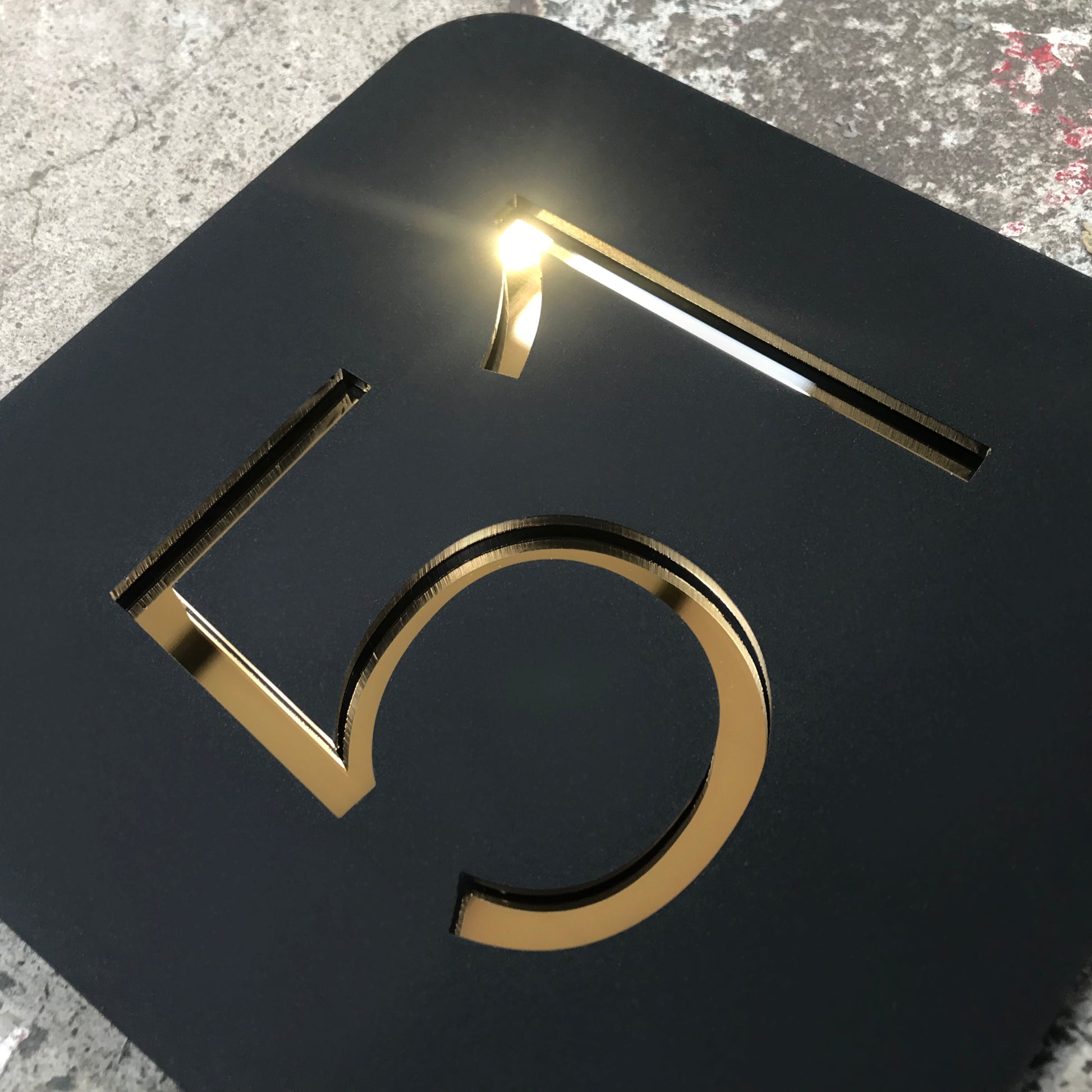 A Stellito rounded square house sign. Comprised of Matt Black Acrylic Face with house number laser cut out, the material is backed with a highly reflective gold mirror, making the numbers sparkle as light hits the numbers.