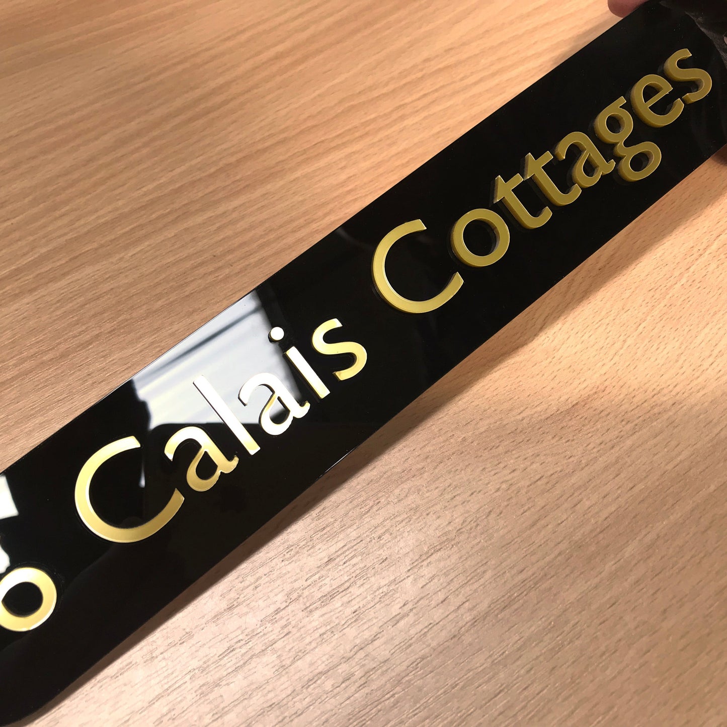 A contemporary and smart looking glossy black sign designed for installation to wooden gates. It depicts a property name 'calais cottages' in rich looking metallic gold acrylic raised letters.